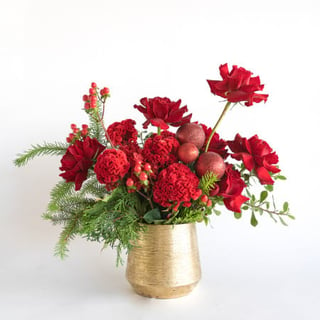 Red roses, celosia, hypericum and baubles arranged with fresh pine in a gold ceramic pot.