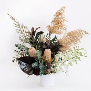 Native flower arrangement featuring banksia, reed or pampass, white orchids, tropical leaves and gum in ceramic vase.