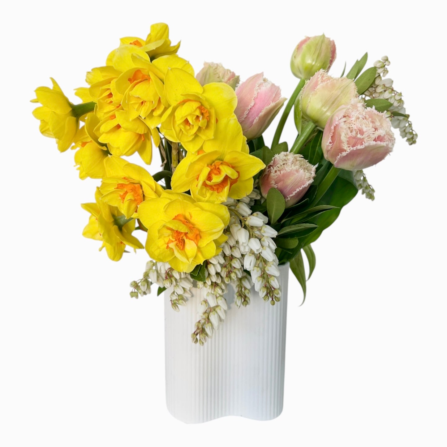 A spring flower bouquet in a white ribbed vase, featuring bright yellow daffodils, pink tulips, and small white bell-shaped flowers, set against a plain white background.