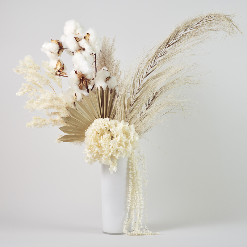 Tall white glass vase featuring preserved flowers with white hydrangea, dried flowers, palm, cotton flower, ming, pampass and amaranthus.