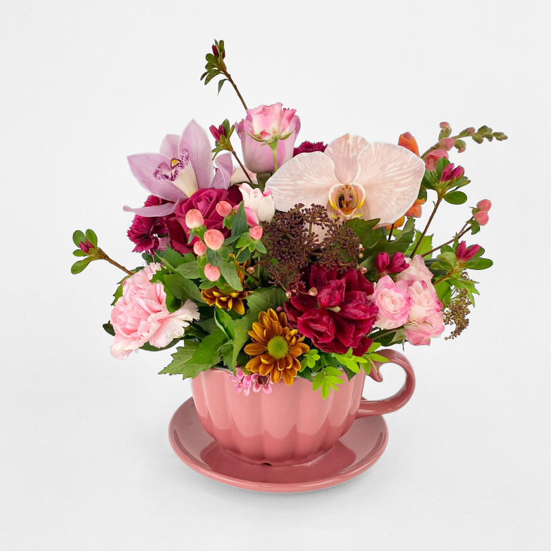 Rose pink teacup and saucer arrangement featuring roses, orchids, carnations and seasonals (varies) to compliment.