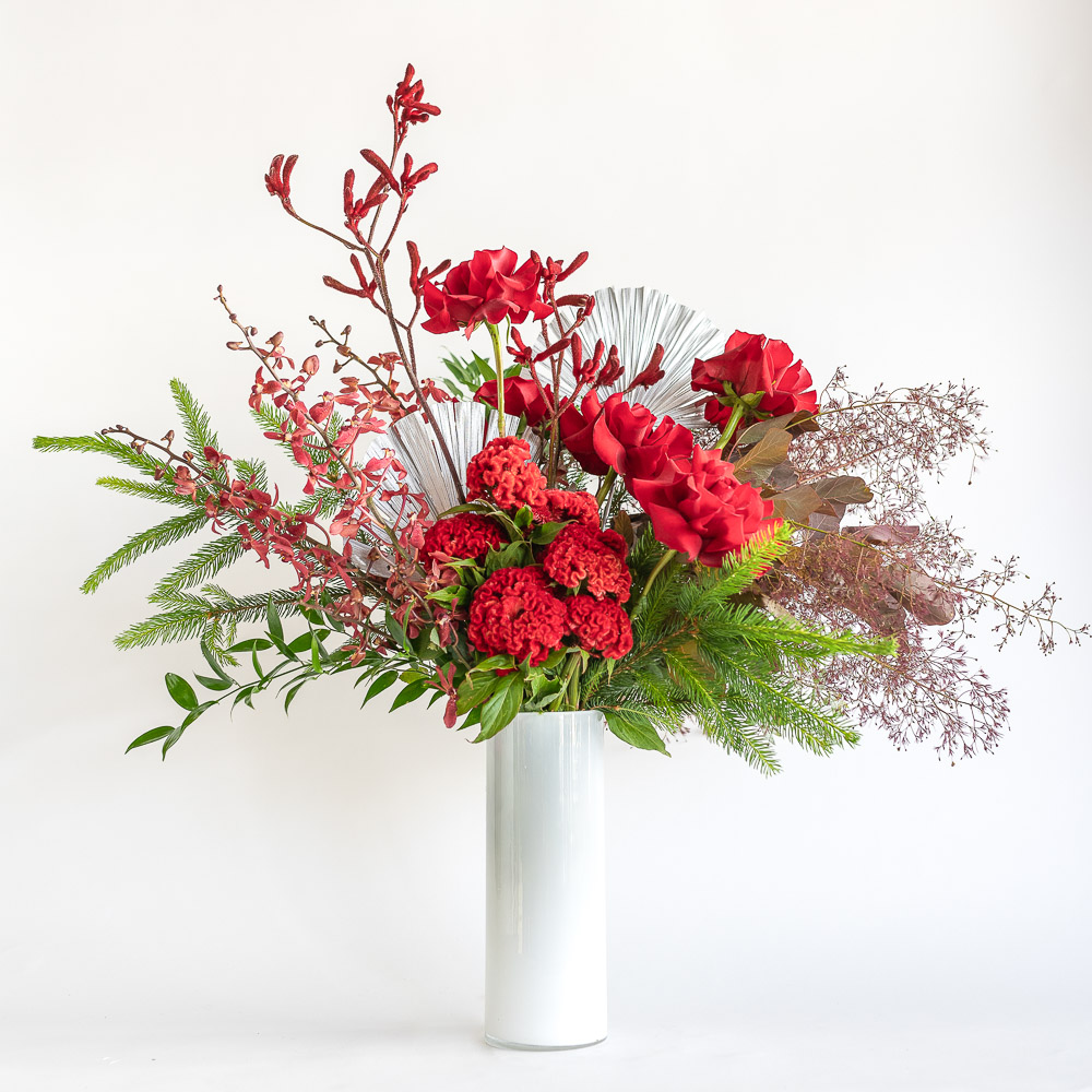 White vase arrangement featuring red roses, celosia, kangaroo paw, dried palm, smokebush, pine, ajumah orchids.