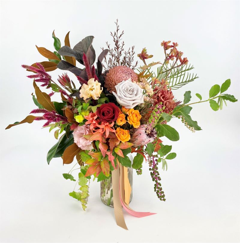 Autumnal themed flowers including roses, protea, disbud, lisianthus and seasonals arranged in a clear vase.