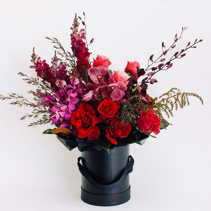 Black hatbox featuring roses, orchids, snapdragons, seasonals to suit with lush dark foliage.